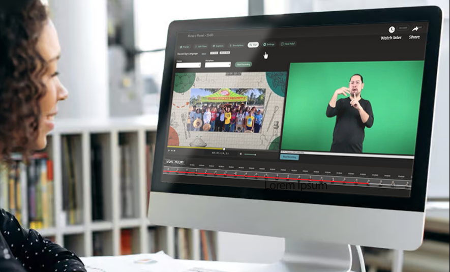 A person sits at a desk using a computer displaying an accessibility-editing interface. The screen shows a split view of a video timeline, a group scene on the left, and an ASL interpreter in front of a green screen on the right. Editing controls and playback tools are visible across the top and bottom of the interface.